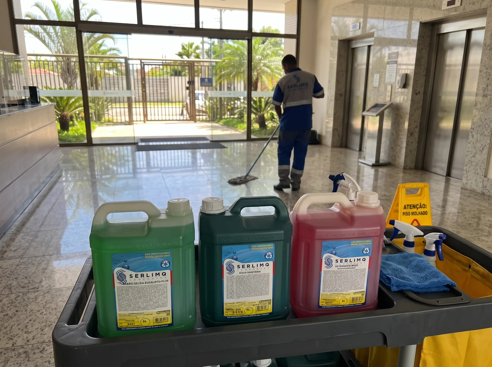 Produtos de limpeza Serlimq em carrinho, com profissional limpando o chão de um saguão moderno